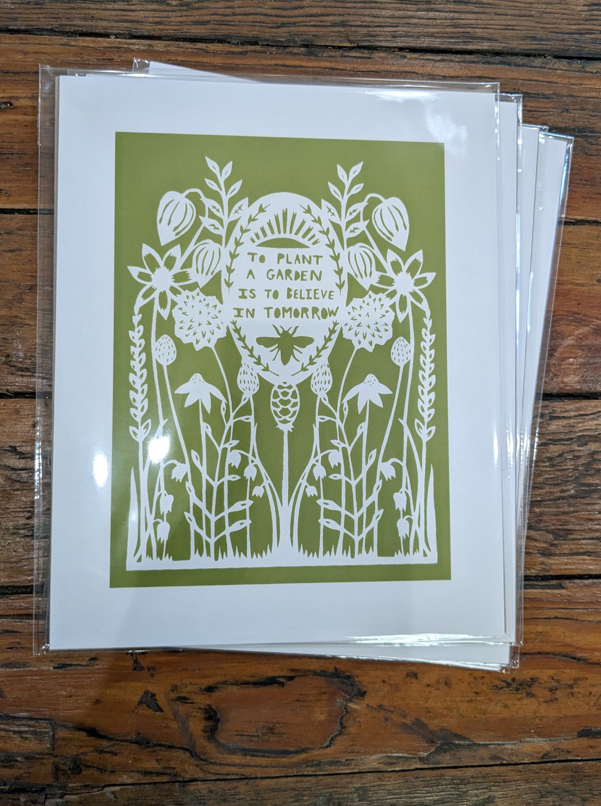 White papercut print on green background depicting a garden: "To plant a garden is to believe in tomorrow"