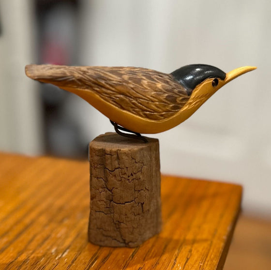 Wood carving depicting a yellow-breasted, black-headed bird with a curious expression, perched on an upright log